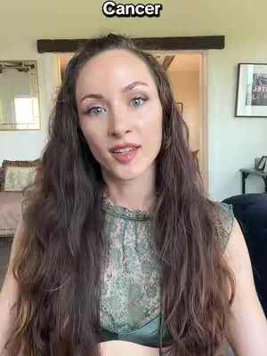 A woman with long brown hair looks straight at the camera, with the word "Cancer" overlaid in white text above her head. She wears a green top, and the background suggests an indoor setting. The overall composition is a selfie style.