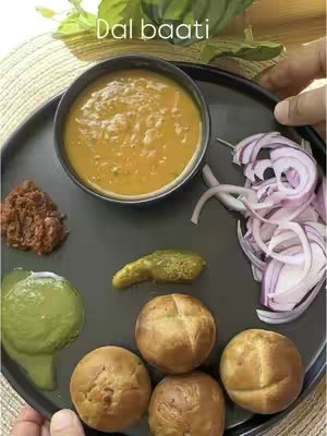 A top-down shot showcases a dark plate filled with various components of a meal. In the center, a small black bowl holds a portion of thick, orange dal. To the left, a blob of chutney and a green relish are visible. On the right, sliced red onions are arranged. Below, four golden, round baatis are arranged in a staggered arrangement.