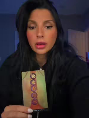 A woman with long dark hair is holding a tarot card in front of her. She's looking at the camera with a neutral expression. The card has a colorful design. The background is softly lit.