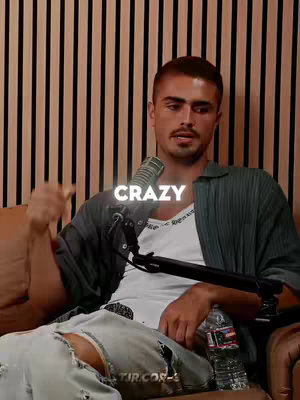 A young man with short brown hair, a neatly trimmed mustache, and a white tank top under a loose, dark-green, button-down shirt is seated. He is positioned in front of a wooden panel background. Above his head, a text overlay reads "CRAZY" in white, outlined text. The man is holding a silver microphone clipped to his shirt and has a water bottle and other items in the foreground. He is speaking towards the camera.