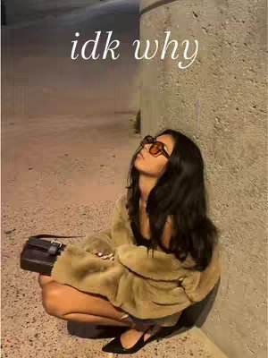 A woman with long dark hair and sunglasses is squatting on the floor, looking up. She's wearing a coat and holding a purse with a sign overlaying the image saying 'idk why'.