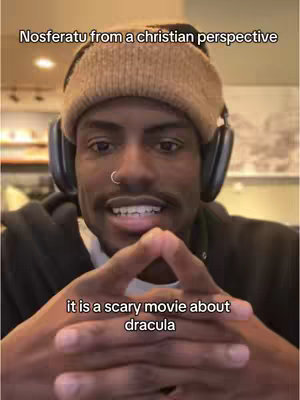A man with a dark complexion and a brown beanie sits in front of a plain background, wearing headphones and making a gesture with his hands. Text overlays describe the image. The lighting is natural and the overall composition is simple.