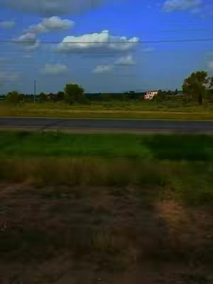 A landscape shot taken from a slightly elevated perspective. The foreground features a grassy area with darker shading at the bottom. The middle ground includes a road, some green fields and distant trees under a bright blue sky filled with puffy white clouds. A building is visible in the distance.