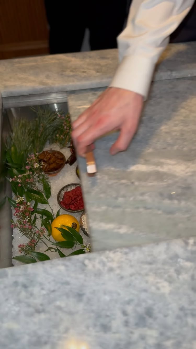 The video shows a hand placing a wooden accessory on a marble surface, where various ingredients are visibly displayed. The hand belongs to a person wearing a light-colored shirt, and some black attire are visible the background. The marble surface is a light grey with veining, and the ingredients are displayed on top of ice inside of a basin. These ingredients include a cut yellow lemon, sprigs of green foliage with small, pink flowers, small bowls with brown and red food displayed.