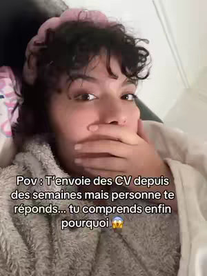 A close-up shot of a person with dark hair and a pink scrunchie covering their head, covering their mouth with their hand and looking at the camera. The person is wearing a fuzzy beige top and is lying down, with text overlayed on the screen.