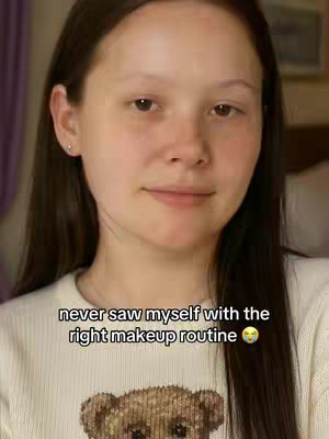 A woman with brown hair and a neutral expression looks at the camera. She's wearing a cream-colored sweater with a teddy bear design in the bottom center. The background is blurred, and text is overlaid at the bottom. It seems like a selfie-style photo, possibly a before photo.