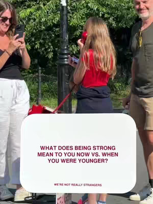The image captures a moment in an outdoor setting. A young girl is holding a red telephone receiver to her ear, seemingly engaged in a conversation. She is seen from the back, wearing a red top, and her blonde hair is flowing down her back. To her left, a woman is holding a phone and taking photo. To the right, a man is standing next to the girl. In front of the girl is a text overlay with the question, "WHAT DOES BEING STRONG MEAN TO YOU NOW VS. WHEN YOU WERE YOUNGER?" The overall scene creates a feeling of candidness and reflection on the theme of strength over time.