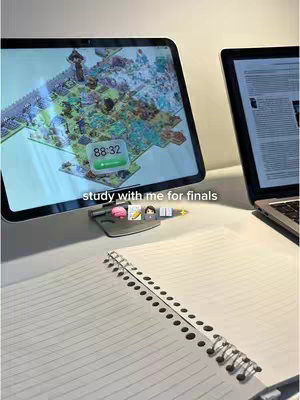 A top-down view of a study setup on a white surface. An open notebook with spiral binding is in the foreground. To the right is an open laptop displaying text, and to the left is an iPad showing a colorful map, with a timer overlay. Below the iPad is a gray stand with various stickers. The top is slightly out of focus, with natural lighting.