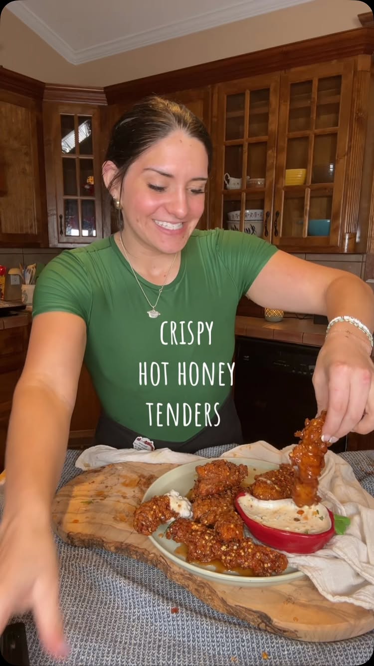 A woman wearing a green top smiles and holds a piece of fried chicken. The image features text overlayed. The background includes wood-paneled cabinets.