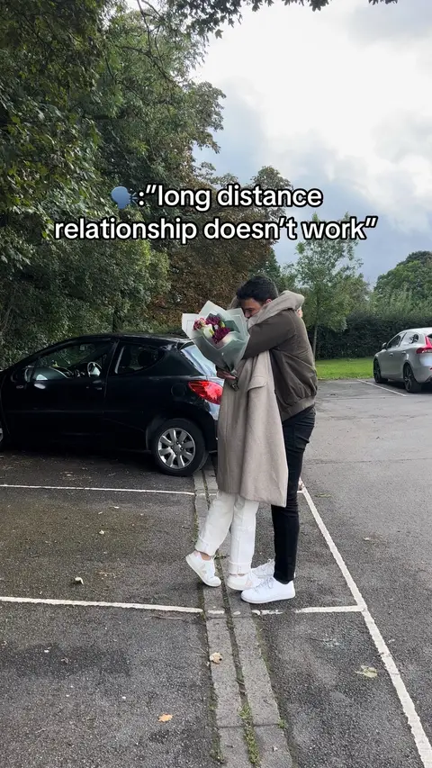 A couple embracing in a parking lot. The man, in a brown jacket, holds a bouquet. Text overlayed at the top. A black car is parked behind them.
