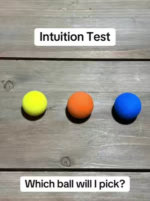 The image is a top-down view of three colored balls—yellow, orange, and blue—arranged in a row on a wooden surface. Above them, the text "Intuition Test" is displayed in a rectangular white box. Below, the question "Which ball will I pick?" is shown on a white box as well, both with simple black font.