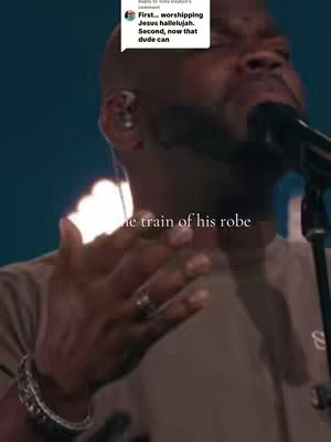 A medium close-up captures a Black man with a shaved head singing into a microphone. He wears a brown shirt, a silver bracelet, and a ring. His expression is focused, and his hand is raised. In the top left corner, a white box contains text.