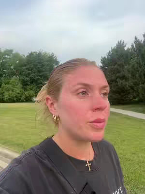 The cover image shows a woman in a natural outdoor setting. She appears to be speaking, with her mouth slightly open. She is wearing a black t-shirt and a gold cross necklace. Her hair is pulled back, and she has gold hoop earrings. The background includes green trees and a cloudy sky. The overall composition is a medium close-up shot.