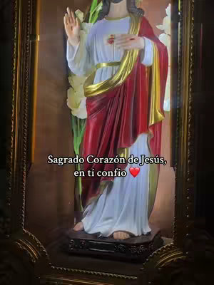 The image shows a statue of Jesus Christ inside a decorative enclosure. The statue is of Jesus in a white robe, a red cloth draped over his shoulders, and a gold sash. He has his left hand over his heart, with a golden heart with a cross on it, and is raising his right hand in a gesture of blessing. The background is dark, with some foliage. Overlayed text reads "Sagrado Corazon de Jesús, en ti confio ❤️".