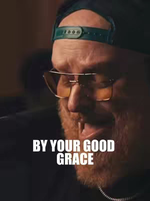 A close-up shot of a man facing left, with a hat and glasses. The man is looking down and slightly to the left side of the frame, and his mouth is open as if he is speaking or singing. The background is a dark, blurred space, ensuring the focus is on the man.
