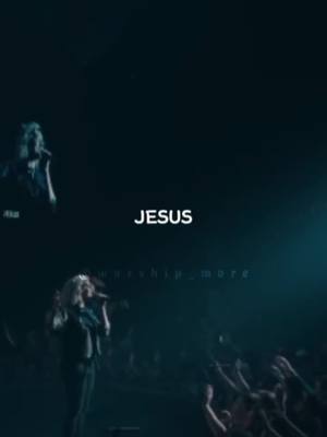 The image is a concert setting dominated by a dark blue color palette. The text 'JESUS' is prominently displayed in white, centered near the top. In the foreground, a woman with blonde hair, wearing a black top, is singing into a microphone, her arms raised. A crowd of people with raised arms fills the lower right corner. In the upper left, a singer is partially visible. The lighting is dramatic, highlighting the performers and the 'JESUS' text.