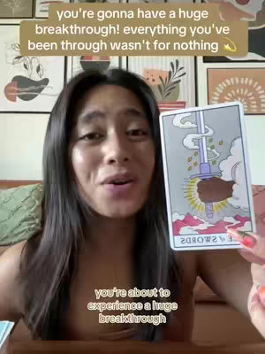 A woman with long dark hair is holding up a tarot card. She appears in casual clothing, looking towards the camera with a slight smile.