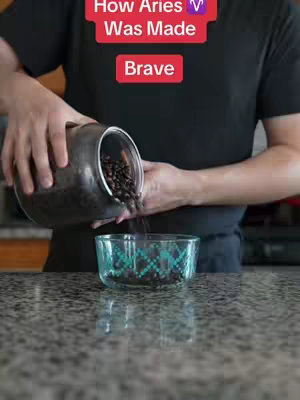 A person wearing a black shirt is pouring dark beans from a glass jar into a clear bowl with a teal pattern, the overall composition is presented at eye-level.