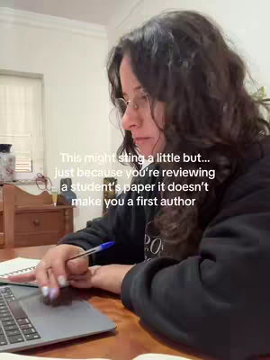 A woman with dark, curly hair is shown from the side, slightly angled toward the camera. She's wearing glasses and a black sweater, and is sitting near a laptop. Her hand is holding a pen, and she's looking down, deep in thought. The background is blurred, but it looks like a home setting with warm lighting.