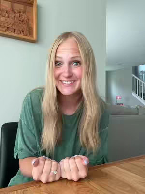 A woman with long blonde hair smiles widely at the camera, her hands clasped together near a wooden table. She wears a green shirt and a ring is visible on her finger. The background suggests a home setting, with glimpses of furniture and a staircase.