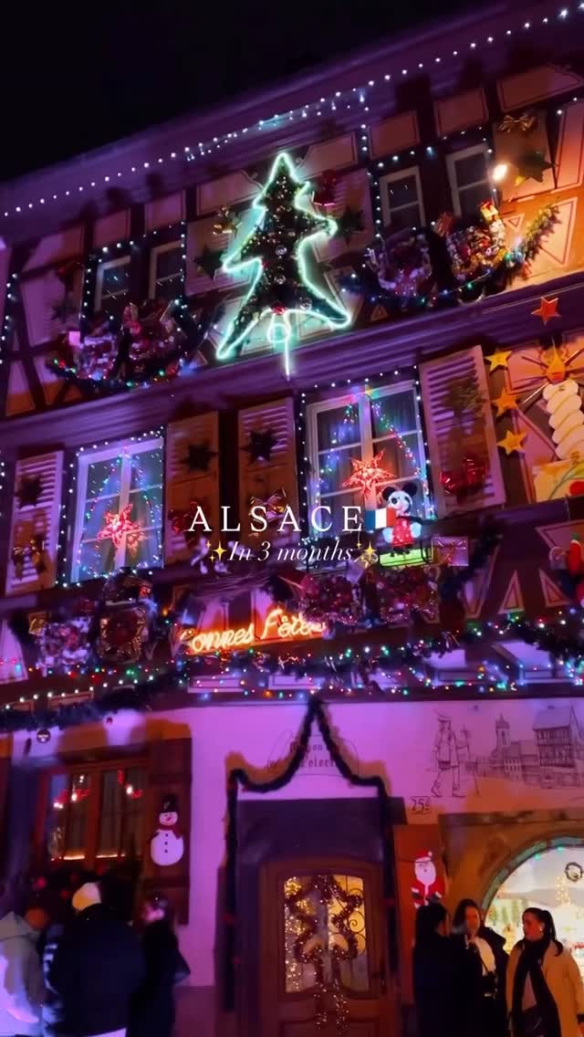 The cover image showcases a beautifully decorated building illuminated for Christmas. The building is a traditional half-timbered structure. Bright lights, garlands, and ornaments adorn the facade, including a large neon Christmas tree. The words 'ALSACE' and 'In 3 months' are overlaid on the image, near the middle. People are visible in the bottom portion of the image, suggesting a street scene.
