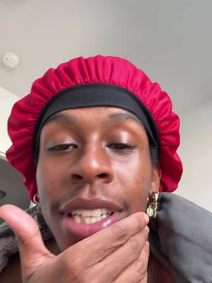 A selfie-style shot focusing on a person's face. The person, who appears to be Black,  is wearing a red bonnet and gold earrings with a green stone. They are looking at the camera with a slight smile, hand near their mouth and chin.