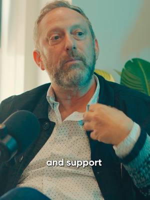 A mid-shot of a man with a graying beard and short salt-and-pepper hair looking off to the side. He wears a white collared shirt under a dark blazer with a green, teal, and blue patterned cuff on his left arm. A black microphone and a green plant are visible to the left and to the right, respectively and the overlaid text "and support" is at the bottom of the frame.