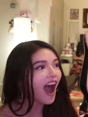 A woman with long, dark hair is the central focus, her widened eyes and open mouth expressing surprise or excitement. The background is a softly blurred interior space, with glimpses of furniture and decor, suggesting a living room setting.