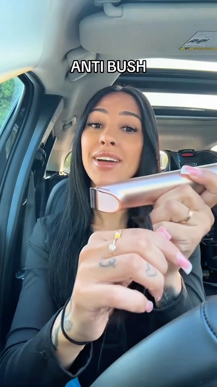 A woman in a car holds a silver electric razor, smiling. The background shows the car's interior, the sun roof, and partially visible trees outside. Text overlay says 'ANTI BUSH'.