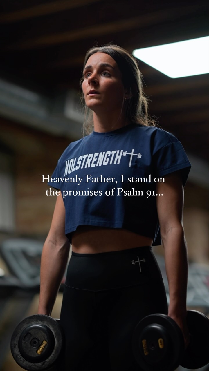 A woman in athletic wear looks upwards with a focused expression while holding dumbbells in a dimly lit setting with gym equipment visible in the background. Text overlayed reads a religious message.
