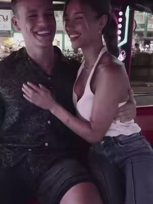 A couple is embracing, laughing, and looking very happy in an outdoor setting, bathed in warm lighting. The woman at the front has her arm around the man's shoulder.