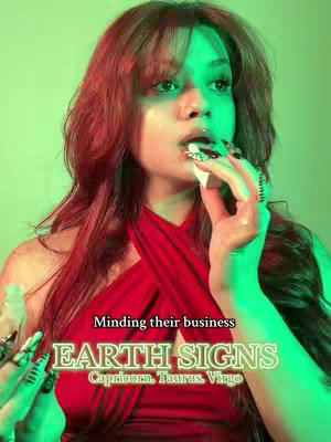 A woman with long, wavy auburn hair stands in front of a green and purple background. She is wearing a halter-style red dress and is holding a white object near her mouth. Overlaid on the image is a text overlay, in a block style, highlighting "Earth Signs" and related zodiac attributes.