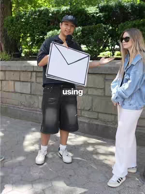 A man in a black hat holding a large white envelope and presenting it to a woman, both standing on a city street, with greenery behind them.