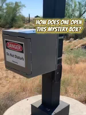 A close-up shot of a gray metal box mounted on a pole. The box has a red and white 'DANGER Do Not Disturb' sticker and is in an environment with dry grass and bushes. The video is likely a puzzle or geocache type.