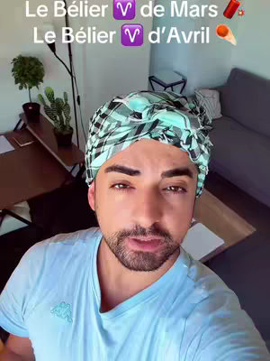 A selfie-style shot featuring a man in his late 20s or early 30s, facing the camera from a medium distance. He's placed centrally, wearing a light blue V-neck t-shirt printed with a logo. A patterned headscarf in shades of blue, green, and gray adorns his head. His skin tone is light. Behind him, a living room setting is visible, including a sofa, potted plants, a desk, and a microphone stand, with soft natural lighting. The color scheme appears to be natural.