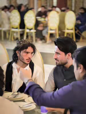 The image captures a social gathering in a restaurant setting, with a focus on three men in a close-up shot. The foreground features a table with plates and silverware, suggesting a meal or dining context. The lighting appears warm, casting soft shadows and highlighting the skin tones of the individuals. In the background, other guests are partially visible, blurred by the depth of field, indicating that the primary focus is on the group in the foreground. Two of the men are looking at each other, while the third man in the foreground is making a gesture, possibly speaking; The overall impression suggests a candid moment in mid-conversation, with an emphasis on interpersonal connection and interactions.
