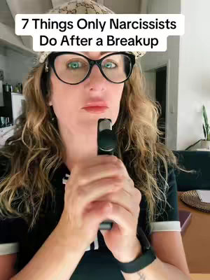 A woman with long wavy hair, wearing glasses and posing. She looks directly into the camera, holding a vape or similar object near her mouth. The background is blurred, and includes neutral-colored wall and white ceiling.