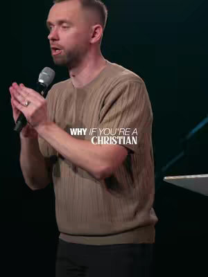 A man in a brown sweater is speaking into a microphone. The background is dark, and text is overlaid on the image.