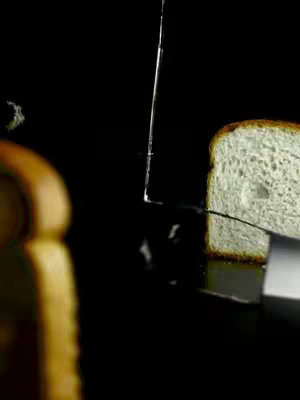 A close-up shot of a slice of bread being cut with a knife against a black background. The bread is rectangular with a light crust and a white interior, and the knife is slicing through. A second slice of bread is in the bottom left corner, and some small bread pieces are slightly visible on the background. The scene is brightly lit, highlighting the bread's texture.
