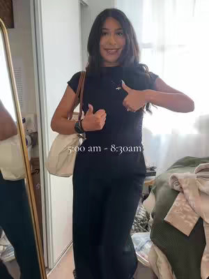 A young woman stands in a bedroom in front of a mirror, giving a thumbs up. The background is a lightly colored, cluttered bedroom with a bed and a closet visible. The woman has dark hair, is wearing a black top and pants and a beige shoulder bag.