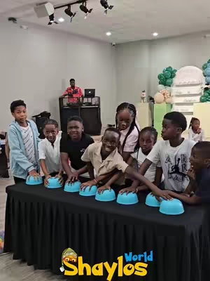 A group of children stand behind a table covered with a black cloth, on which are several blue, dome-shaped objects. They appear to be interacting with these objects, possibly playing a game, in a brightly lit indoor space. A DJ is in the background.