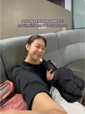 A young woman with brown hair is seated on a gray sofa, holding a blue passport and smiling. The image is a selfie, with the camera held at arm's length. Overlayed text at the top reads, "NYC INTERNSHIP DIARIES: moving into my NYC apartment." The overall composition is casual and warmly lit.