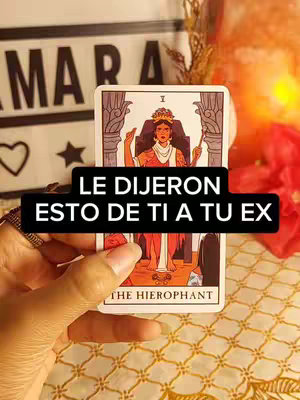 A hand is holding a tarot card with the The Hierophant, with a black text box overlaid reading "LE DIJERON ESTO DE TI A TU EX". Behind the card is a blurred background including what appear to be some other decorations, and the card is in front of a patterned cloth.
