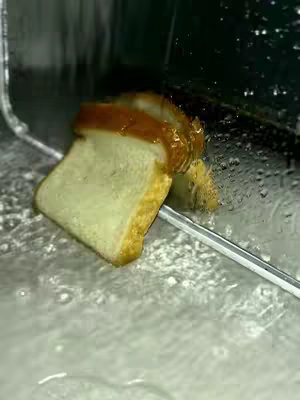 A slice of bread, appears to be submerged in water or a watery substance, possibly with jam on top. The bread leans towards the right as if being affected by the medium. The composition features an interplay of transparency and reflection, possibly shot underwater in a sink.