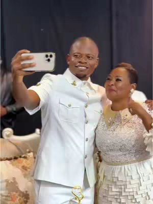 A selfie-style photo featuring a man taking the photo while standing next to a woman. Both are smiling and dressed in formal, light-colored attire. The man is holding the phone up with his right hand at arm's length, positioning himself for the shot. The woman stands beside him. The background is blurred and dark.