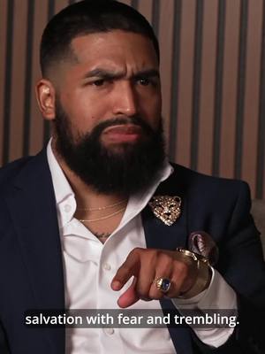 A man with a beard and wearing a suit jacket is looking at the camera with a serious expression. He is pointing his index finger towards the viewer. The background is an out-of-focus wall with vertical stripes.