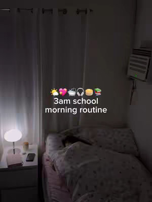 The cover image shows a dimly lit bedroom with the bed in focus. A person is under the covers. On a nightstand next to the bed is a lamp, a small container, a phone, and a pen. The hook text "3am school morning routine" is overlaid on the image, with relevant emojis