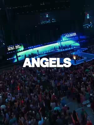 An overhead shot of a large outdoor concert. The stage is lit with blue and green lights, and a large crowd of people is visible in the foreground. The word "ANGELS" is prominently displayed in white text over the crowd. The sky is dark, suggesting it is night.