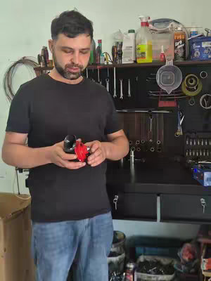 A man holding two mechanical parts in a workshop-like setting, with various tools and supplies in the background