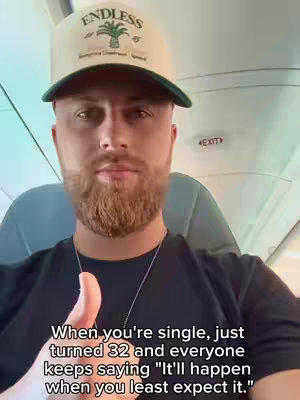 A selfie-style video of a man in an airplane wearing a baseball cap. He is looking at the camera and giving a thumbs up. Text below the man provides a caption about being single.